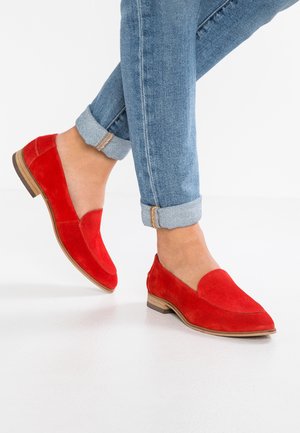 Person wearing bright red suede loafers with wooden soles paired with rolled-up light blue denim jeans on a white floor.