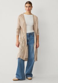 Cardigan lungo beige con apertura frontale, polsini a coste, abbinato a jeans a zampa leggeri blu chiaro e scarpe beige senza lacci. Maglietta bianca semplice sotto.