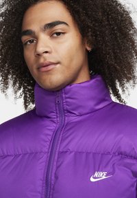 Giacca puffer Nike viola con colletto alto, zip frontale e logo bianco sul lato sinistro del petto. Tessuto liscio, design leggero.
