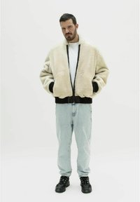 Beige faux faux pels bomberjakke med sorte kanter og manchetter, båret over en hvid T-shirt, parret med lysvaskede jeans og sorte sneakers.