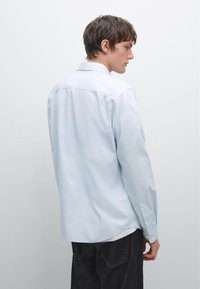 Camicia a maniche lunghe di colore azzurro chiaro con colletto a punta, polsini con bottoni e vestibilità rilassata. Tessuto liscio senza motivi o dettagli visibili.