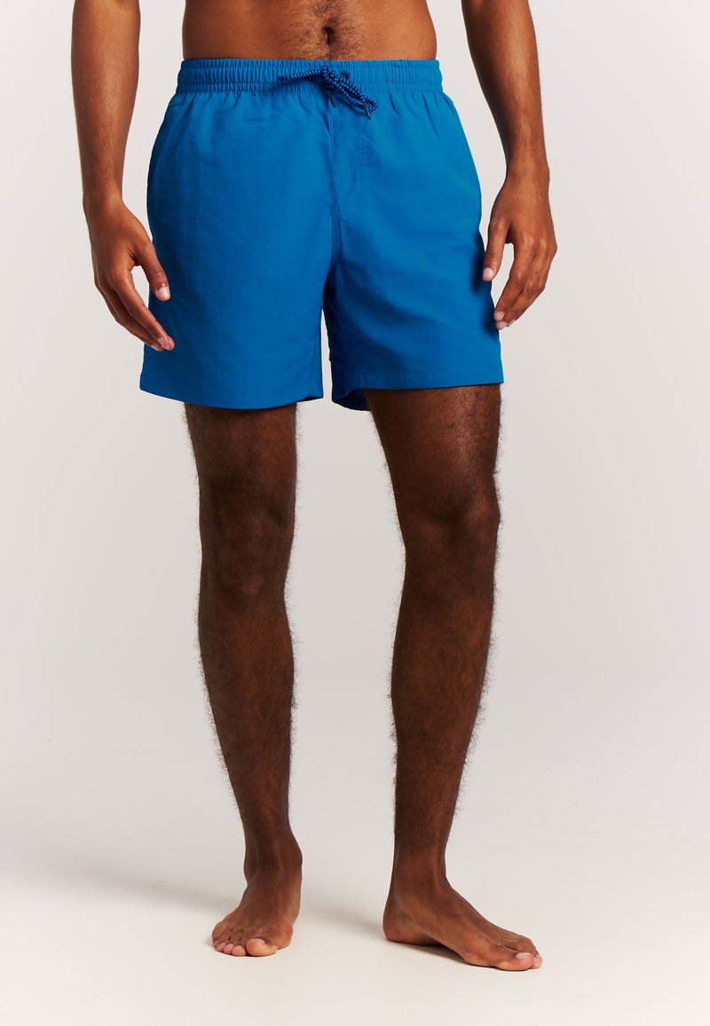 Hombre con pantalones cortos de baño azul brillante de pie descalzo contra un fondo neutro, con piernas y manos visibles.