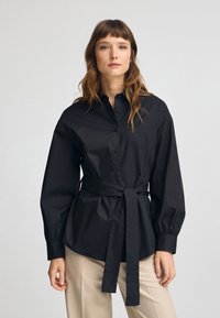 Czarna koszula z guzikami, z wcięciem w talii i długimi bufkami, wykonana z gładkiej bawełny. Posiada usztywniony kołnierz i przewiązany pasek.