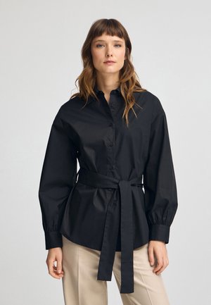 Camisa negra de botón con cintura ceñida y mangas largas abullonadas, hecha de algodón suave. Presenta un cuello estructurado y un cinturón atado.