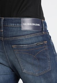 Ciemnoniebieskie dżinsy o gładkim wykończeniu. Posiadają skórzaną łatkę z logo na pasku i charakterystyczne przeszycia tylnej kieszeni.