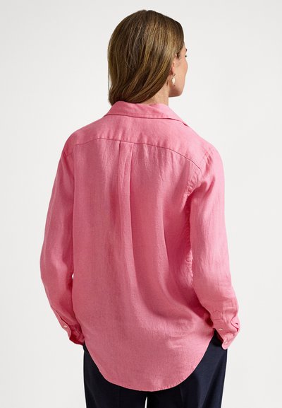 Sieviete ar gaiši brūniem matiem, valkājot vaļīgu rozā garroku kreklu un tumšas bikses, redzama no aizmugures uz balta fona.