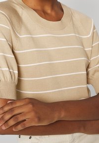 Beige striktrøje med hvide vandrette striber, rund halsudskæring og pufærmer. Blød tekstur, tætsiddende design, synlige ribbede manchetter.