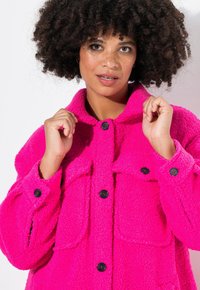 Chaqueta de forro polar en un rosa brillante, con cuello, cierre de botones y dos bolsillos frontales. El tejido texturizado añade interés visual.