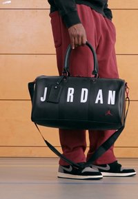 Fekete bőrből készült sporttáska nagy fehér "JORDAN" felirattal, piros Jumpman logóval, két fogantyúval és levehető vállpánttal.