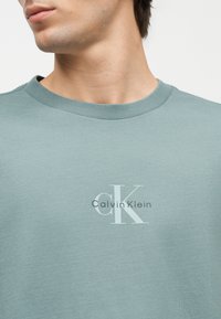 Nærbilde av en person som har på seg en lys grønn T-skjorte med rund hals og en hvit og grå Calvin Klein-logo midt på brystet.