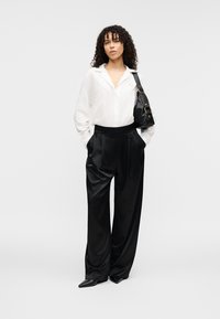 Λευκό πουκάμισο με κουμπιά, μαύρο σατέν παντελόνι φαρδιάς γραμμής, μαύρη τσάντα με χρυσές λεπτομέρειες και μυτερά μαύρα παπούτσια. Μπούκλες στα μαλλιά, ουδέτερο φόντο.