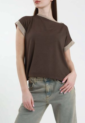Donna che indossa una maglietta marrone scuro a maniche corte e larga con bordo beige e jeans chiari, in piedi con una mano in tasca e l'altra lungo il corpo.