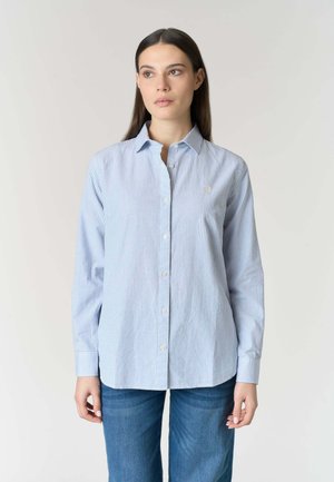 Donna che indossa una camicia a righe sottili azzurre e bianche con maniche lunghe e jeans blu, in piedi davanti a uno sfondo semplice.