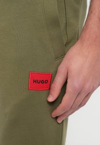 Detailný záber na ruku ležiacu pri vrecku na olivovo zelených nohaviciach s červenou obdĺžnikovou nášivkou s logom "HUGO".