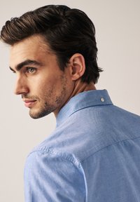 Šviesiai mėlynas mygtukų marškinėlis su struktūrizuota apykakle, tekstūruotu audiniu ir mygtuko akcentu nugaros pusėje. Prisitaikantis dizainas.