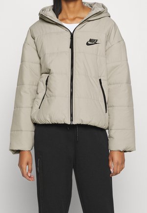 Beige Nike dunjakke med hætte, sort lynlås og lommer, båret over hvid skjorte, parret med sorte bukser med lynlåslomme.