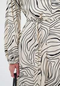 Kermakleid mit schwarzem abstraktem Wellenmuster, langen Ärmeln und elastischen Bündchen. Verfügt über einen gebundenen Taillengürtel für eine verstellbare Passform.
