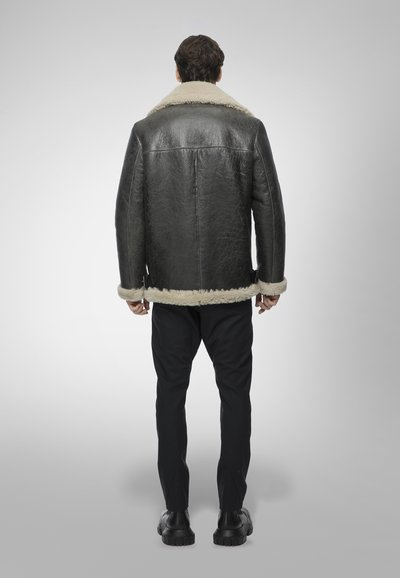 Uomo in piedi, di spalle, che indossa una giacca in shearling nera con bordo in pelliccia color crema, pantaloni neri e scarpe nere su sfondo grigio.