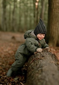 Vaikas apsirengęs žaliu paminkštintu sniego kostiumu atsilenkia ant rąsto, dėvi dryžuotą juodą kepurę ir smėlio spalvos pirštines, supamas rudens lapų ir medžių.