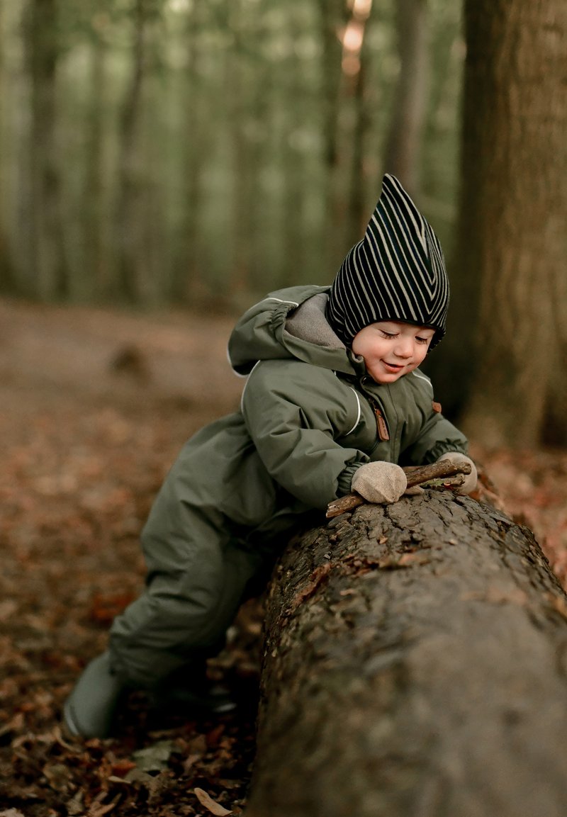 Vaikas apsirengęs žaliu paminkštintu sniego kostiumu atsilenkia ant rąsto, dėvi dryžuotą juodą kepurę ir smėlio spalvos pirštines, supamas rudens lapų ir medžių.
