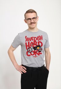 Camiseta gris de manga corta con un gráfico de motocicleta en rojo y negro que incluye el texto "SWEDISH HARD CORE" en tipografía roja en negrita.