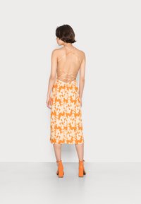 Vestido laranja com estampado floral, com comprimento médio, costas abertas e alças cruzadas. Combinado com sandálias de salto laranja.