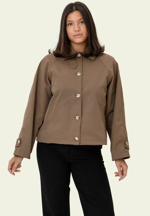 Noor naine sirgete tumedate juustega, seljas nööbitud pruun jakk ja mustad püksid, seisab ühevärvilise heleda tausta ees.