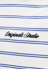 Άσπρο ύφασμα με ομοιόμορφα τοποθετημένες λεπτές μπλε οριζόντιες ρίγες και μαύρο κεκλιμένο κείμενο "Originals Studio" κεντημένο κοντά στο κέντρο.