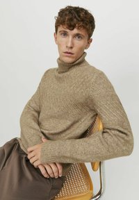 Maglione beige a collo alto lavorato a maglia con motivo a spina di pesce testurizzato. Maniche lunghe, polsini aderenti e colletto a costine. Indossato con pantaloni marroni.