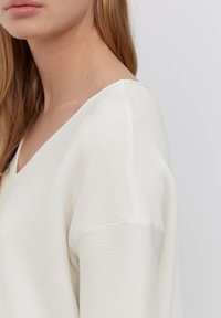 Weißer V-Ausschnitt Pullover mit strukturierter Stoffstruktur, verfügt über eine lockere Passform und abgefallene Schulternähte. Klare Linien und minimale Details.