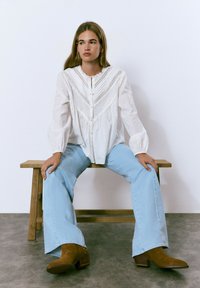 Blusa blanca con tejido texturizado, detalles de encaje y botonadura frontal, combinada con jeans de campana azul claro y botas de tacón marrones. Sentada en un banco de madera.