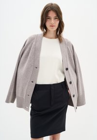 Světlý šedý oversized cardigan s ribbovanými manžetami a lemem, s předním zapínáním na knoflíky, nošený přes krémový top a černou tužkovou sukni. Hladká textura.