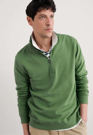 Man wearing green quarter-zip sweater over a white-collared striped shirt, beige pants, looking to the side against a plain background.