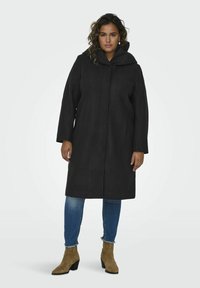 Manteau noir en laine avec capuche, fermeture à boutons sur le devant, manches longues et coupe droite. Associé à un jean bleu et des bottines marron.