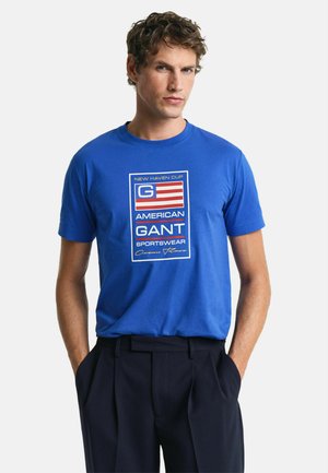 Blue cotton t-shirt featuring a graphic print with red and white stripes, logo text, and a crew neck design. Paired with black trousers.