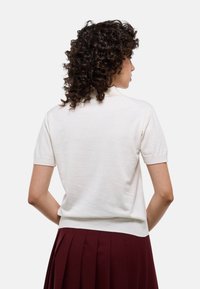 Pull à manches courtes en maille de couleur blanc cassé, avec un col et un ourlet côtelés. Texture douce, silhouette ajustée, porté sur une jupe plissée noire.