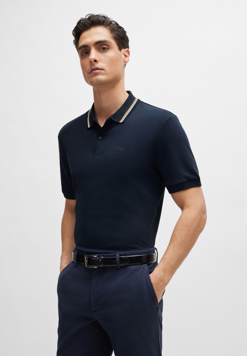 Polo de color azul marino con mangas cortas, cuello con franjas en color crema en contraste y bordado sutil del logo en el pecho. Combinado con pantalones oscuros.