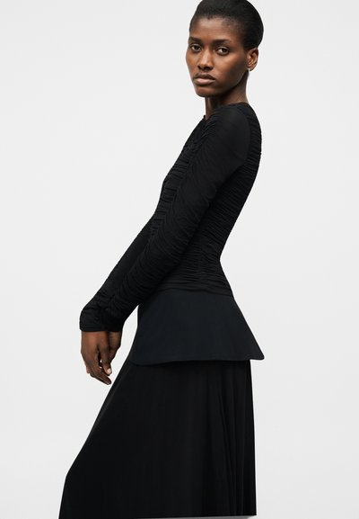 Kvinde med mørk hud iført en tætsiddende sort ruched bluse med lange ærmer og en flydende sort nederdel, poserer mod en ensartet lys baggrund.