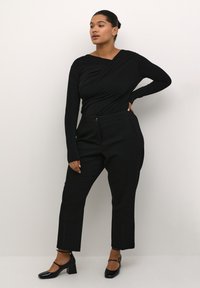 Haut noir à manches longues enroulé avec un décolleté plongeant, associé à un pantalon noir taille haute. Le mannequin porte des chaussures à talons carrés en vernis noir.