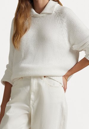 Femme aux longs cheveux portant un pull en maille blanc avec col et un pantalon blanc taille haute, une main reposant sur la hanche.