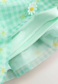 Tela de tul verde menta con un patrón de cuadros, decorada con margaritas blancas bordadas y acentos amarillos. Textura suave y ligera.