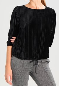 Schwarzes Samtoberteil mit vertikalen Falten, Rundhalsausschnitt und Dreiviertelärmeln. Mit Kordelzugdetails an der Taille, kombiniert mit grauen strukturierten Hosen.