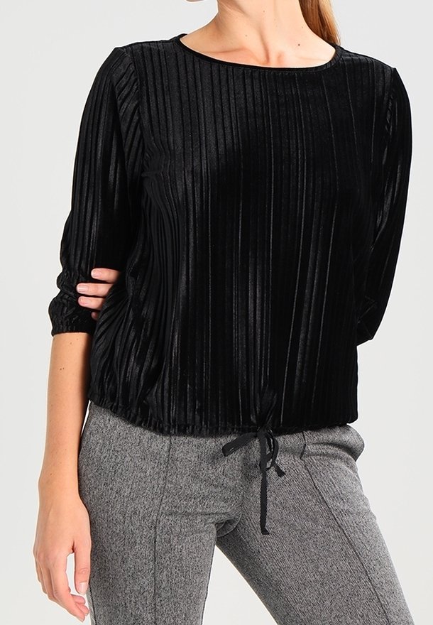 Schwarzes Samtoberteil mit vertikalen Falten, Rundhalsausschnitt und Dreiviertelärmeln. Mit Kordelzugdetails an der Taille, kombiniert mit grauen strukturierten Hosen.