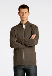Maglione zip con collo alto di un marrone scuro, realizzato in tessuto morbido, con polsini e orlo a coste, indossato sopra una camicia bianca.