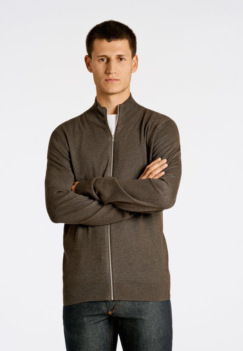 Maglione zip con collo alto di un marrone scuro, realizzato in tessuto morbido, con polsini e orlo a coste, indossato sopra una camicia bianca.