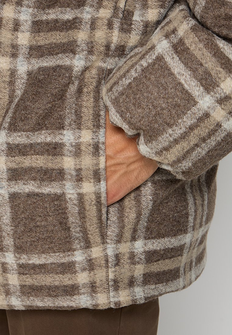 Veste à carreaux marron et beige en tissu doux et texturé, avec une poche latérale et des manches retroussées, mettant en valeur le motif à carreaux.