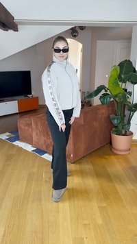Heledhall hallakas zippidega jakk logoriba, mustad püksid ja hallid saapad. Keskkonnas on taim ja tekstuuriga pruun diivan.
