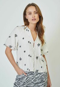 Blusa bianca a maniche corte con ricami floreali neri e chiusura con bottoni. Il tessuto ha una texture stropicciata. Accoppiata con una gonna a righe grigia.