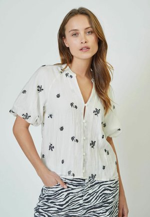 Blusa bianca a maniche corte con ricami floreali neri e chiusura con bottoni. Il tessuto ha una texture stropicciata. Accoppiata con una gonna a righe grigia.