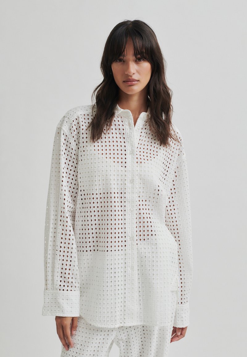 Bela bluza z mrežastim vzorcem, s izrezanimi detajli, dolgimi rokavi in gumbnim zapiranjem. Lahkotna tkanina, primerna za plastenje.